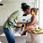 Mother presses her forehead against her daughter’s forehead as her young daughter sits on the kitchen counter.